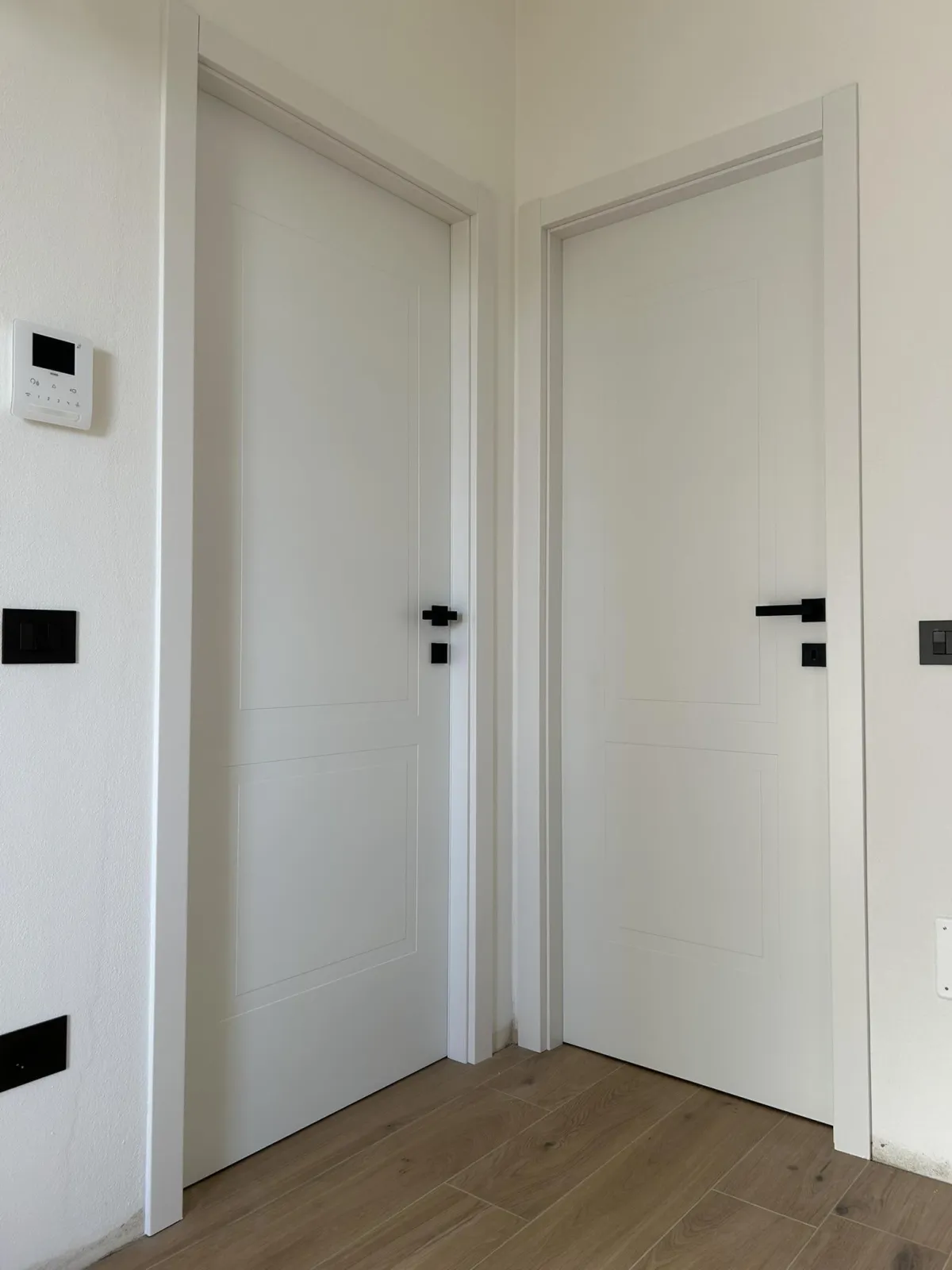 Coppia di porte interne laccate bianche ad angolo con maniglie nere minimaliste a contrasto e pavimento in legno chiaro.