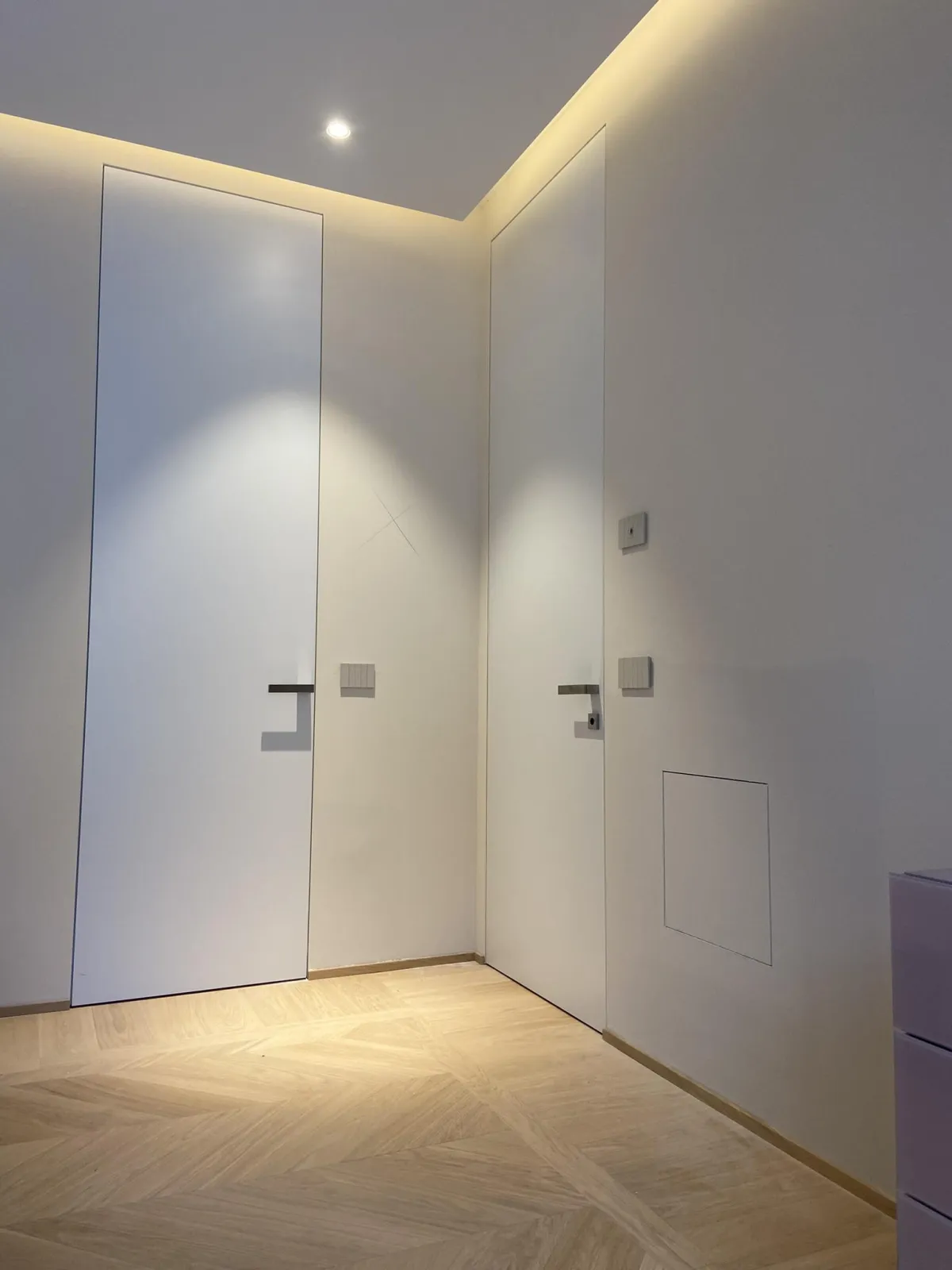 Una fotografia cattura un moderno corridoio interno con pavimento in parquet a spina di pesce chiaro e un'illuminazione a soffitto incassata e calda. Sono visibili due porte interne bianche a filo muro con maniglie minimaliste.