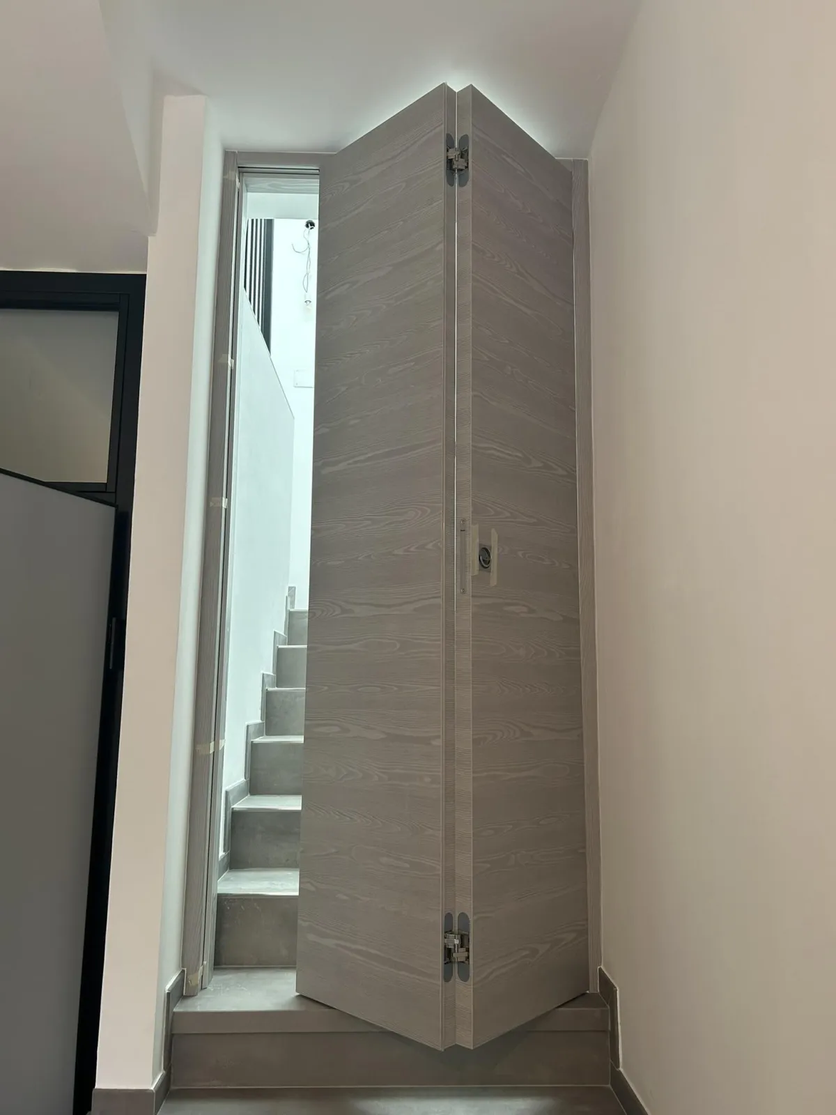 Una fotografia di una porta interna aperta con finitura a grana di legno grigio-marrone, che rivela scale di cemento che conducono a un pianerottolo superiore moderno in uno spazio dalle pareti bianche.
