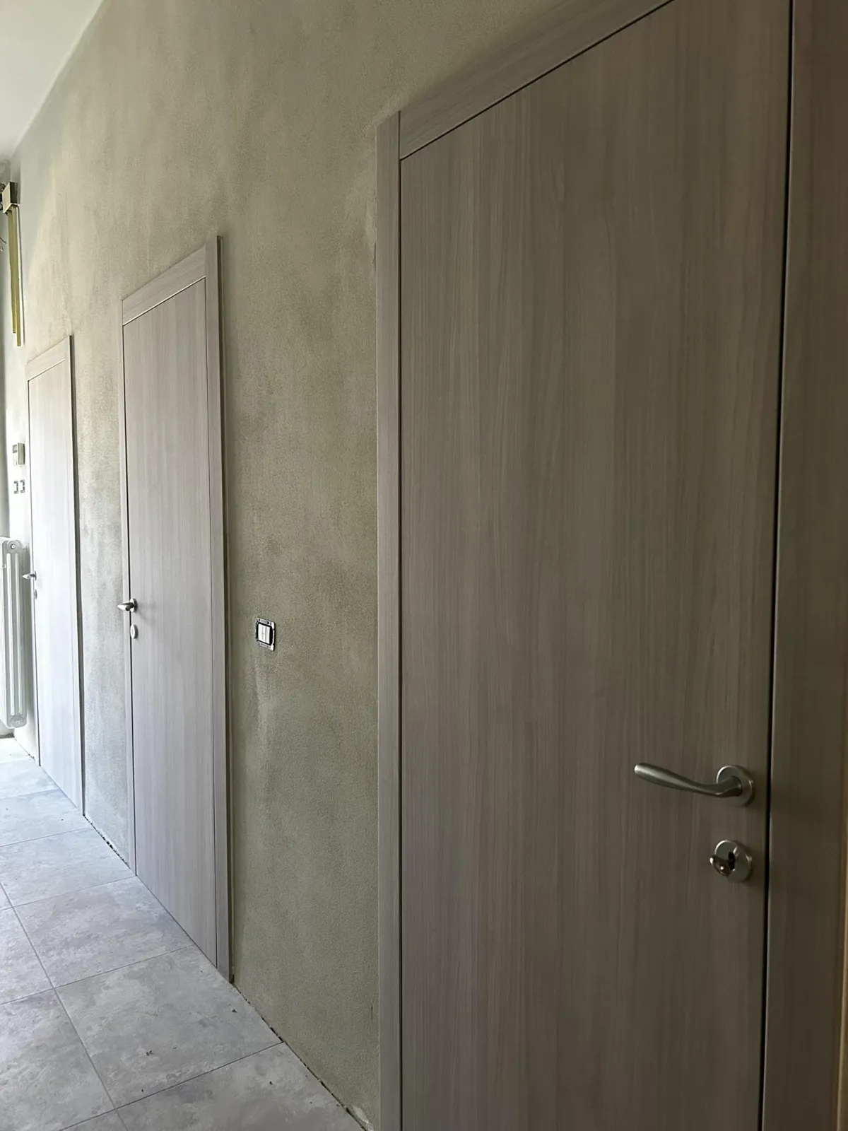 Porte interne moderne in laminato effetto legno grigio chiaro installate in un corridoio con pareti effetto cemento.