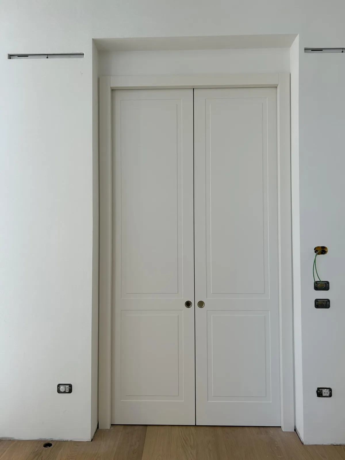 Una fotografia che mostra una coppia chiusa di porte interne a pannelli bianchi moderni incastonate in una parete bianca. Sotto le porte sono visibili diverse prese elettriche e il pavimento in assi di legno chiaro scorre ovunque.